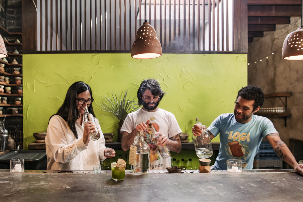 Harlan and her two colleagues take a second to stand behind a bar in front of a green painted wall, smelling mezcal 