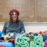 Ephraim Ruttenberg sits at a table covered in crochet creations, a crochet hat on his head, a chalkboard covered with mathematical equations behind him.