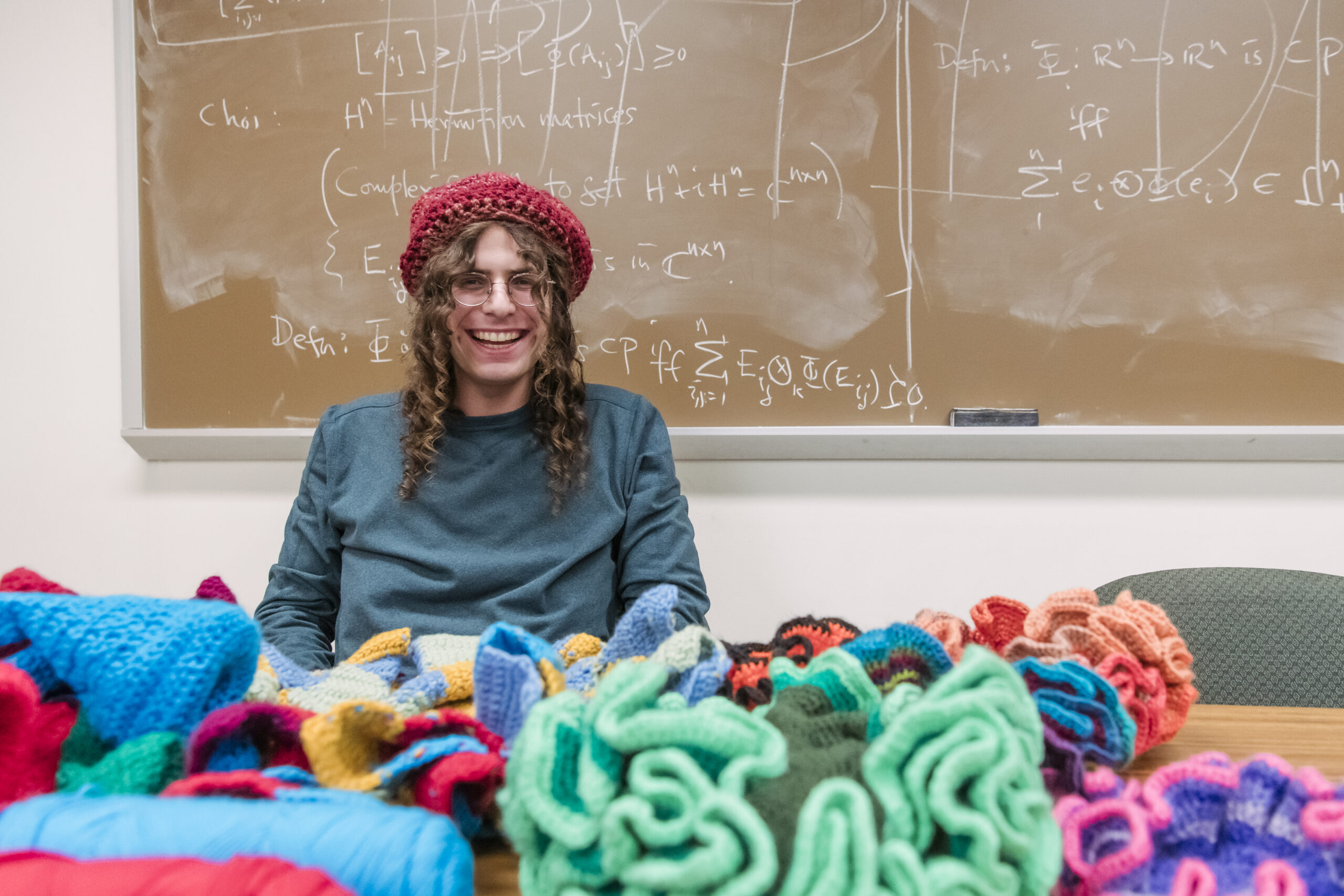 Ephraim Ruttenberg sits at a table covered in crochet creations, a crochet hat on his head, a chalkboard covered with mathematical equations behind him.