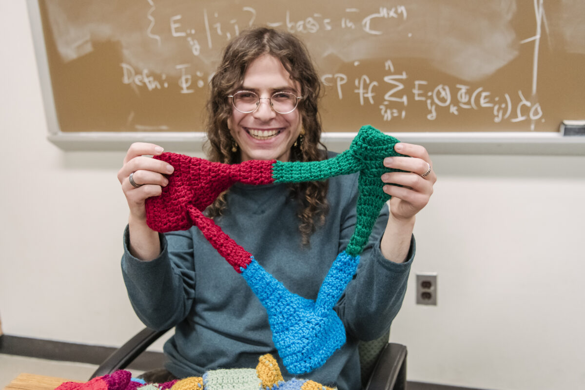 Ephraim Ruttenberg holds up a crochet creation that is roughly an open triangle, with three distinct sections in green, red, and blue; he is backed by a chalkboard with equations