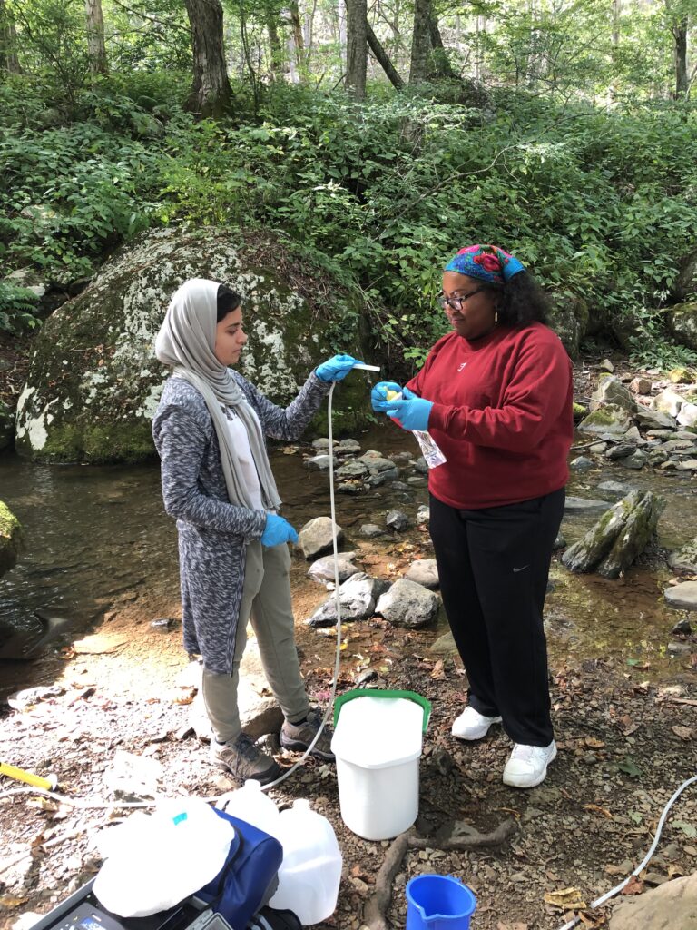 two women stand on the bank of a stream; one holds the end of a long thin tube above a plastic bag held by the other. Some buckets and other equipment are on the ground nearby. 
