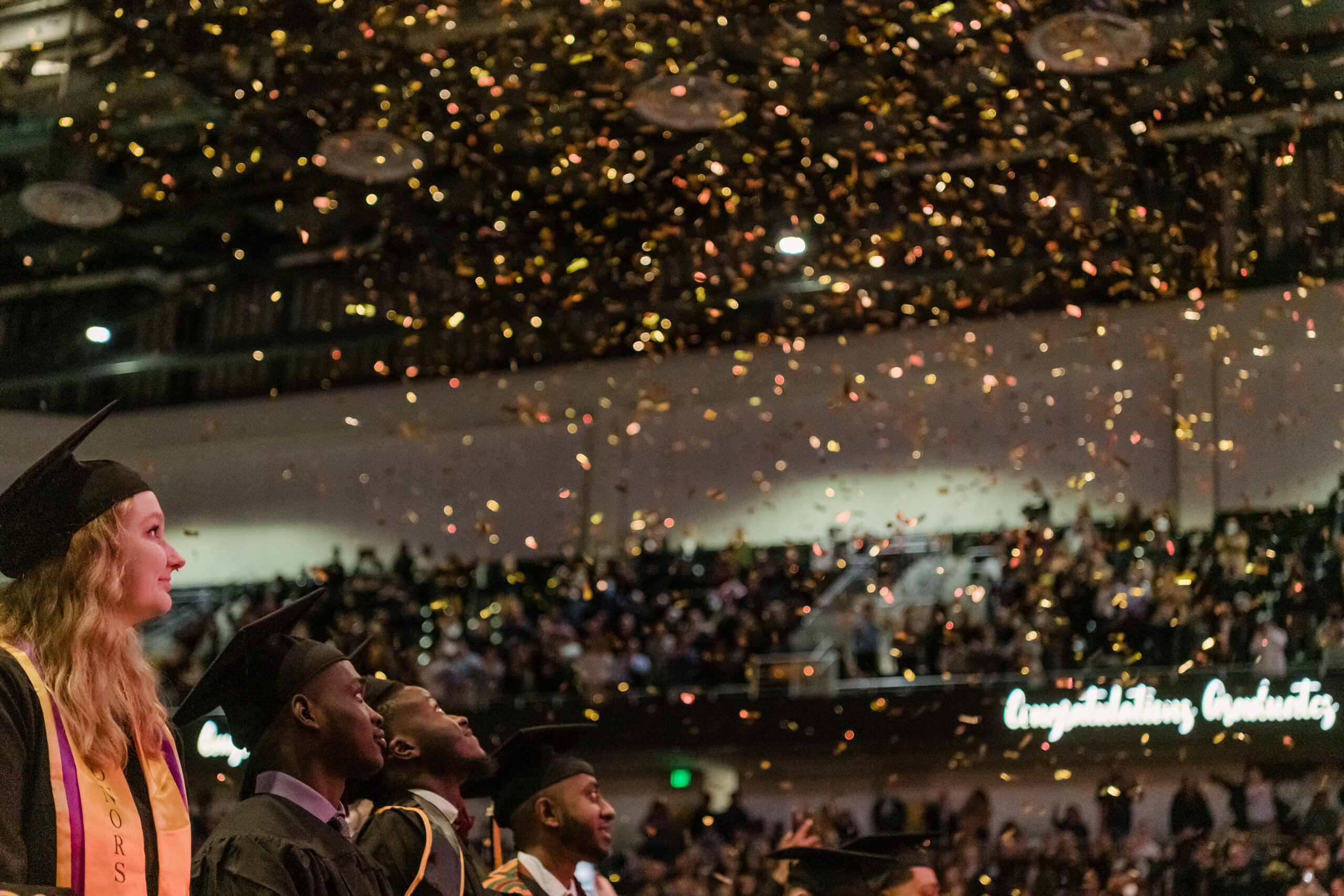 students in commencement regalia look up as glitter falls