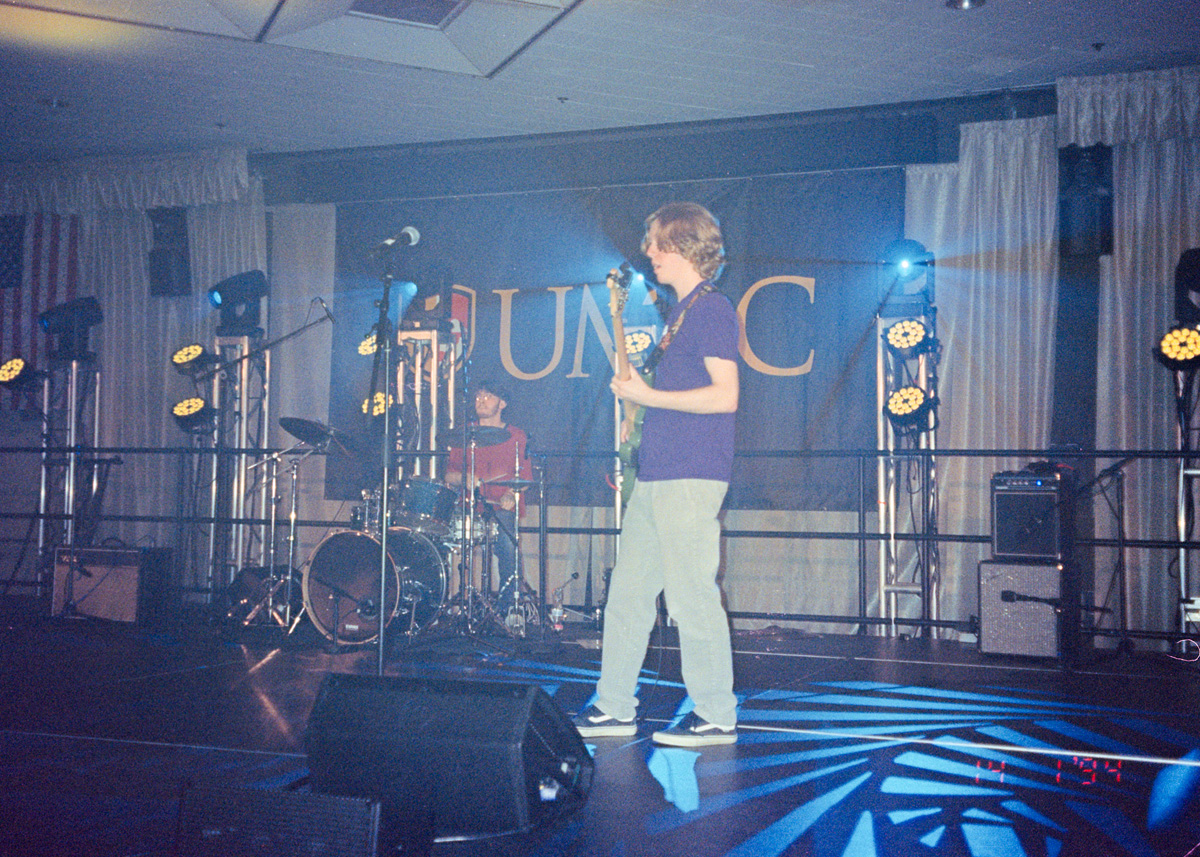 Two students on stage playing music. One standing towards the front playing bass guitar, and the other one in the background playing the drums. Stage lights shine overhead.