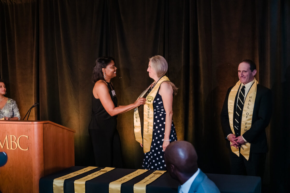 Karen Woodard at the 2022 UMBC Athletics Hall of Fame ceremony placing a commemorative stole around Dana Eberly Keiner, Volleyball, 1993-96.