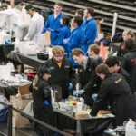 Multiple teams of students wearing lab coats and safety glasses stand around tables. On top of the tables are chemicals, beakers and other lab equipment.