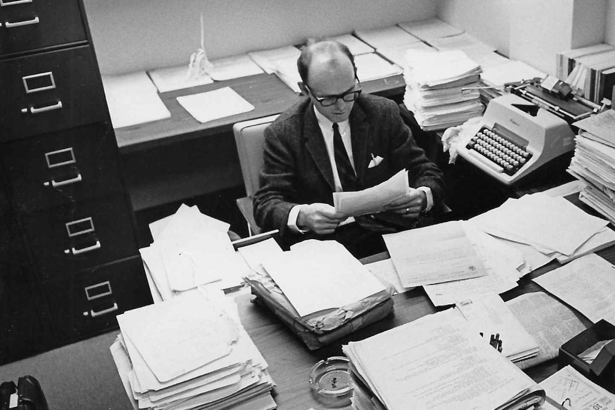 Chuck Peake at his desk in 1969, surrounded by piles of papers.