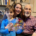 An older man, woman, and young child pose together. The child holds a transparent clock with colorful gears inside.