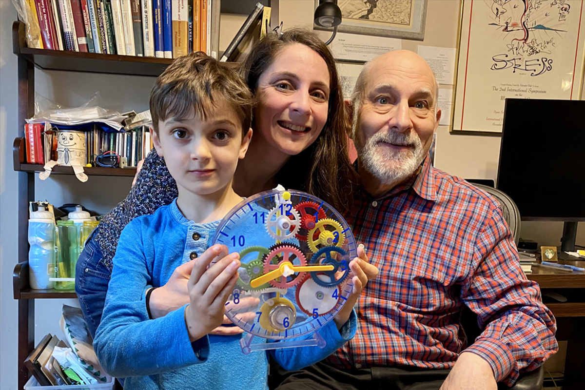 An older man, woman, and young child pose together. The child holds a transparent clock with colorful gears inside.