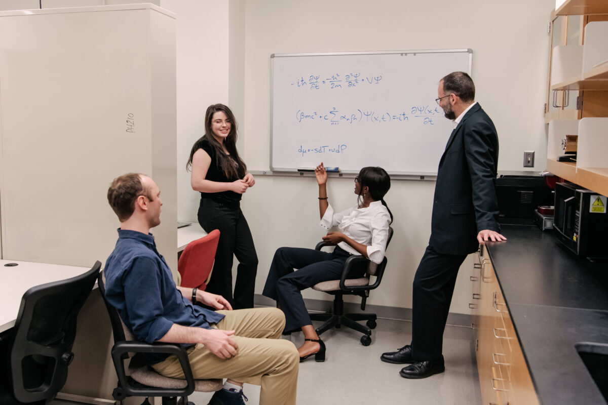 four people, two seated and two standing, discuss thermodynamics equations on a wall whiteboard nearby.