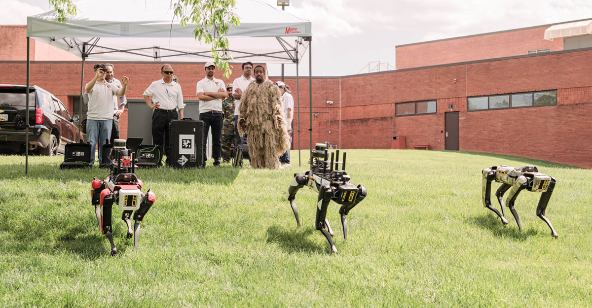 On the back lawn of the courthouse building, student researcher Kevin Rippy, left, controls three Spot robots with gestures. Another student stands by in a ghillie suit, a type of camouflage clothing.