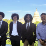 Three college students in business suits stand with a college professor in a balcony with the U.S. Capitol behind them.