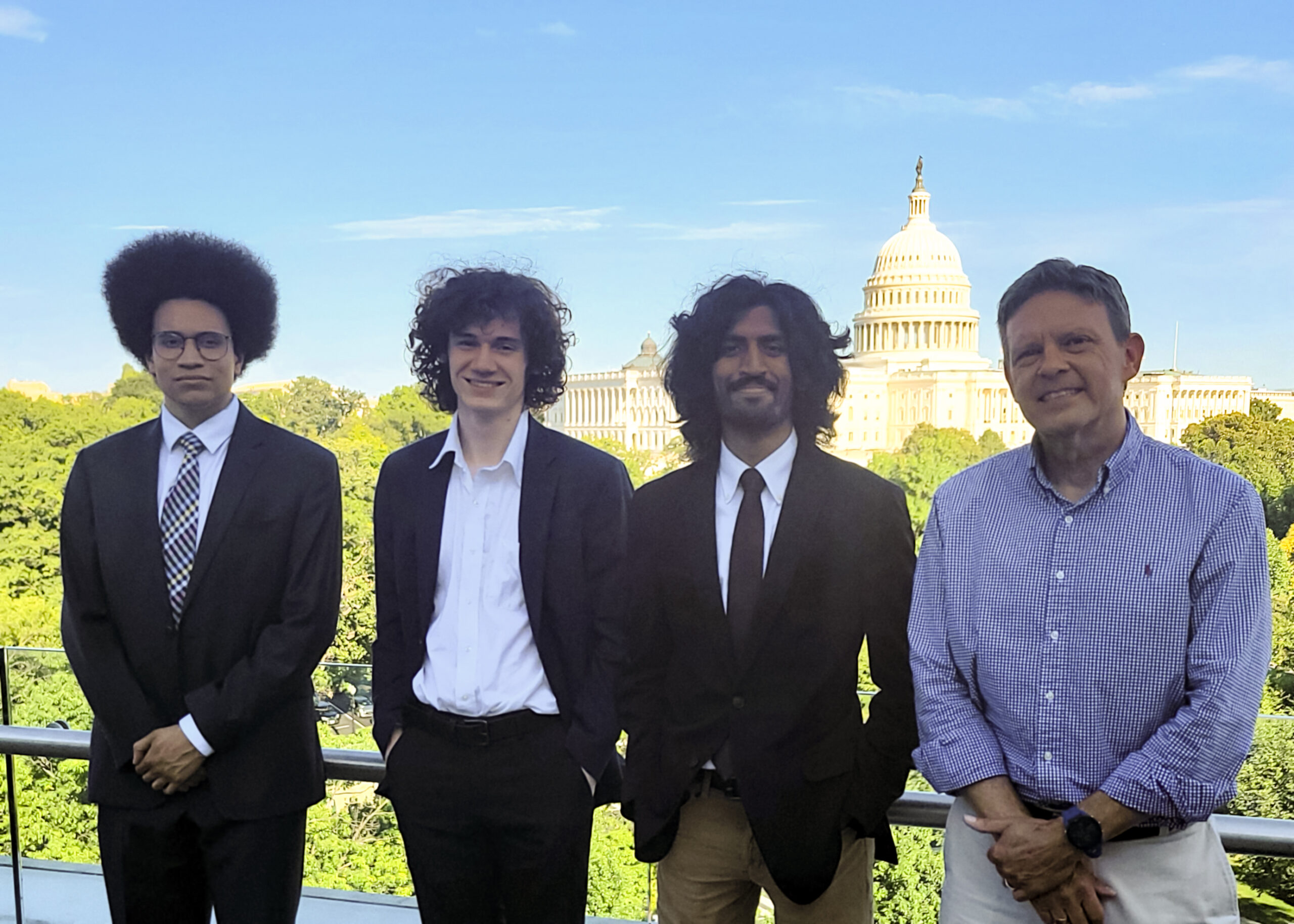 Three college students in business suits stand with a college professor in a balcony with the U.S. Capitol behind them.