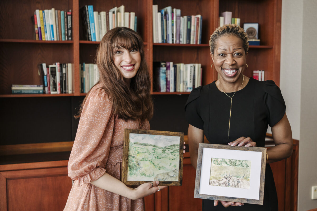 Two women smiling, standing in front of bookshelf, holding scenic landscape paintings.