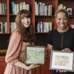 Two women smiling, standing in front of bookshelf, holding scenic landscape paintings.