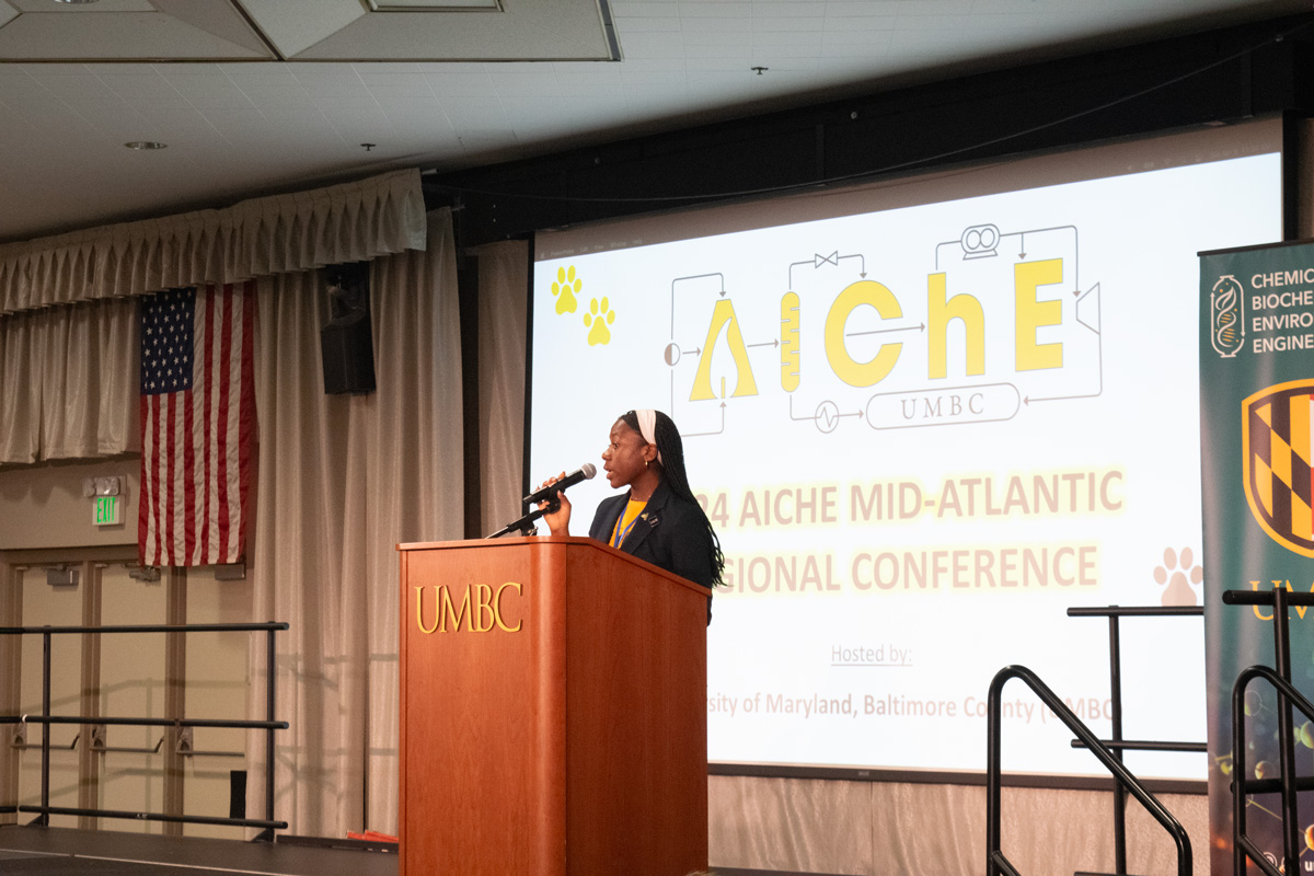 A woman stands at a podium with the letters UMBC on it. In the background is a slide that says AIChE Mid-Atlantic Regional Conference.