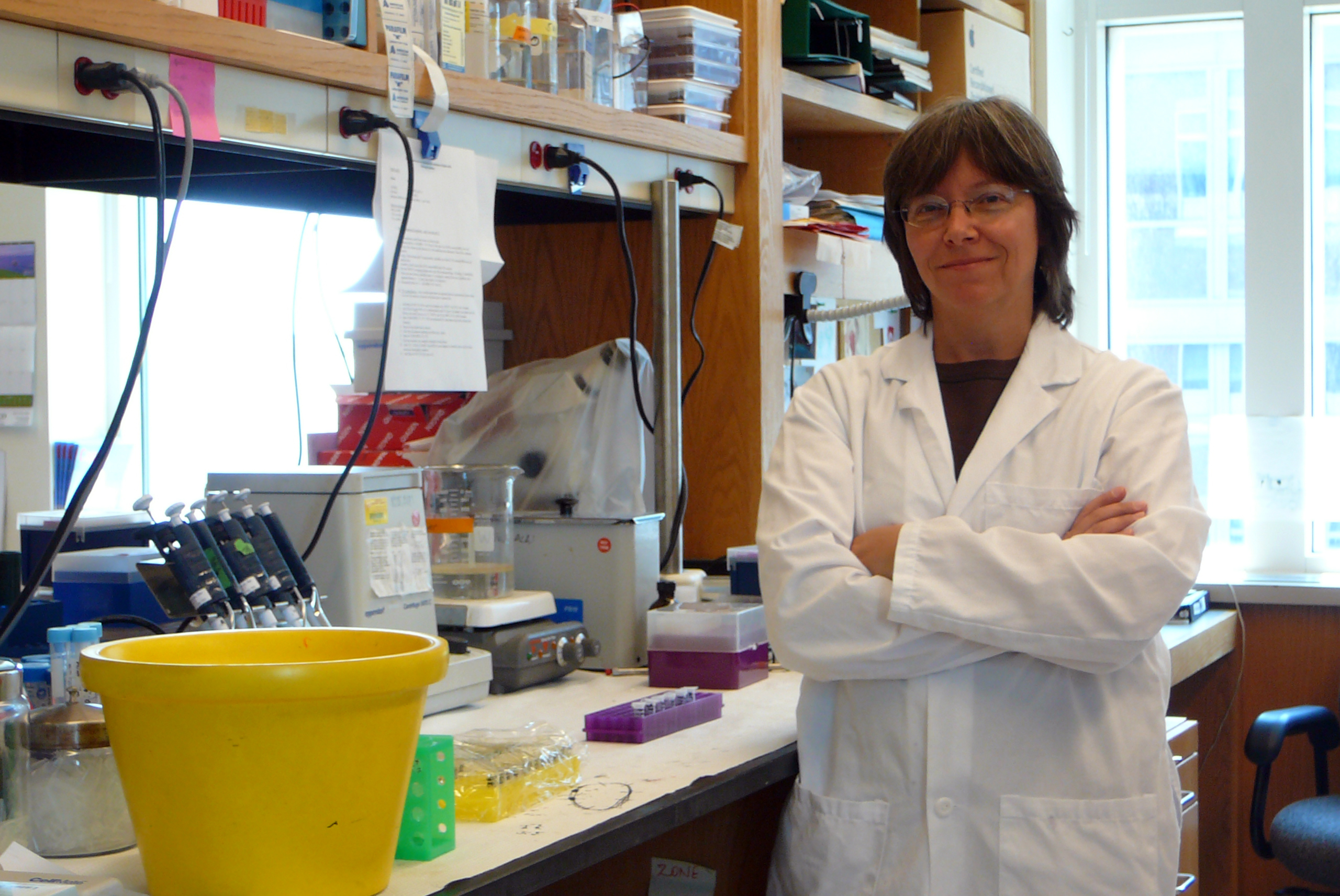 In a laboratory, a woman in a white lab coat and glasses smiles at the camera