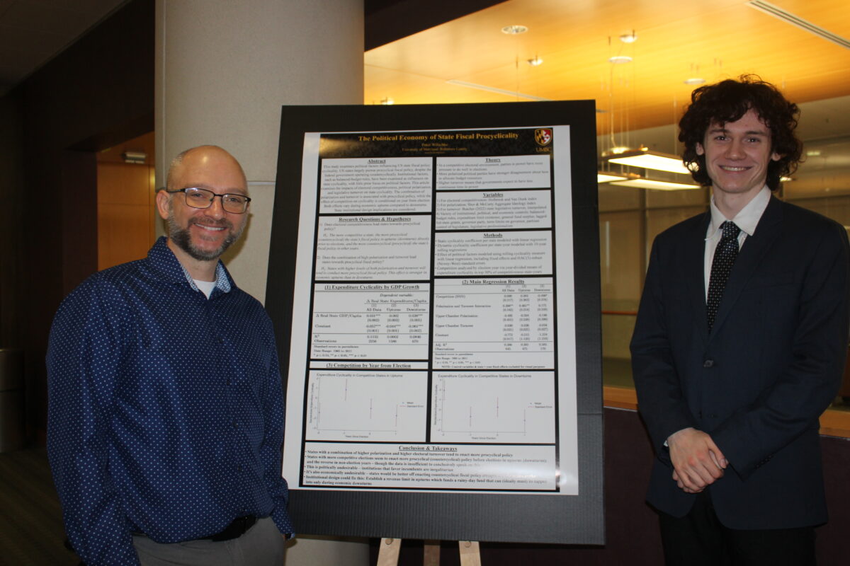 A professor and Peter Wilschke stand on both sides of a research paper set on an easel in a large room with hanging lights and a wood ceiling