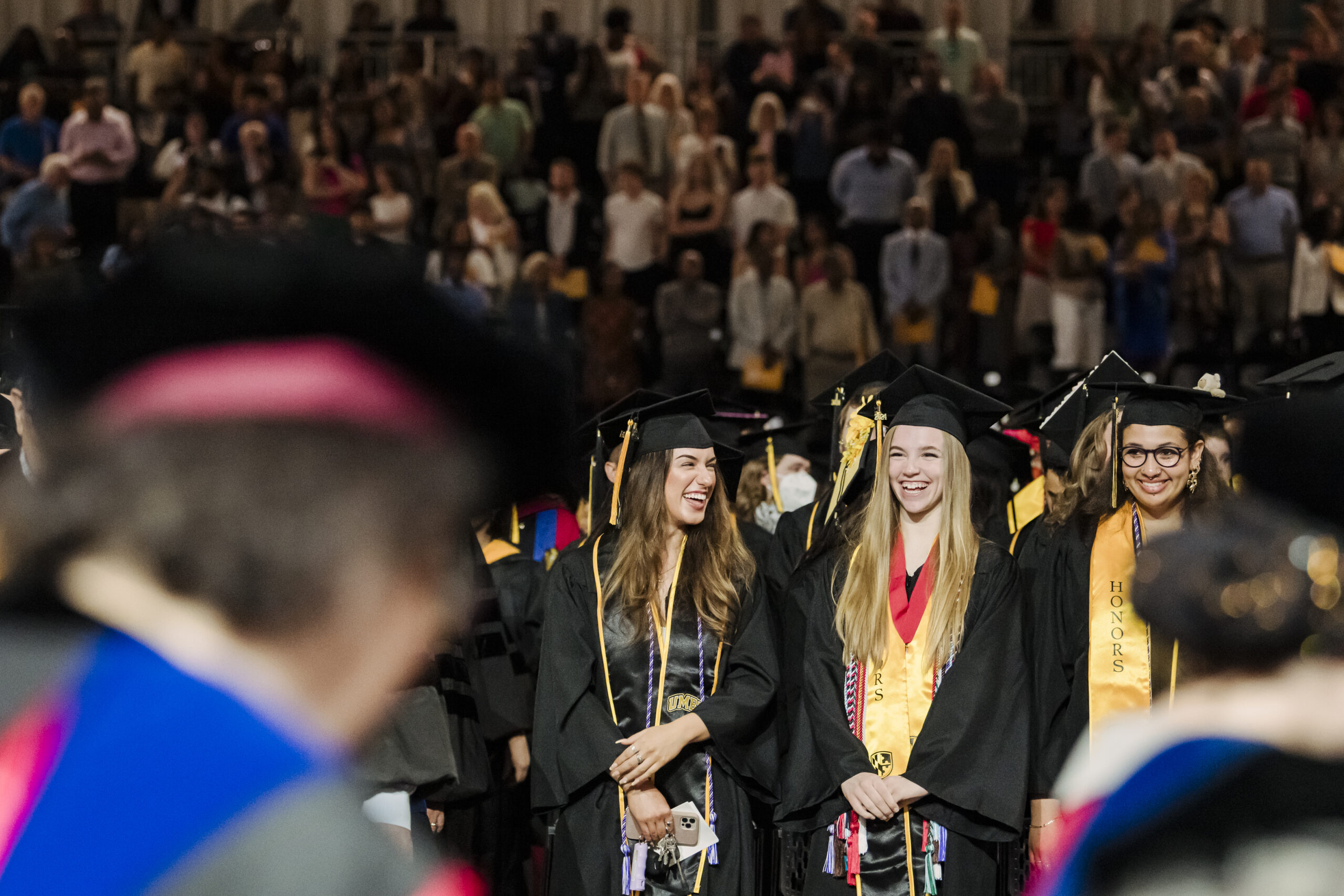 Three UMBC graduates during the Spring 2024 Commencement ceremony. The students are smiling while surrounded by the audience in the background.