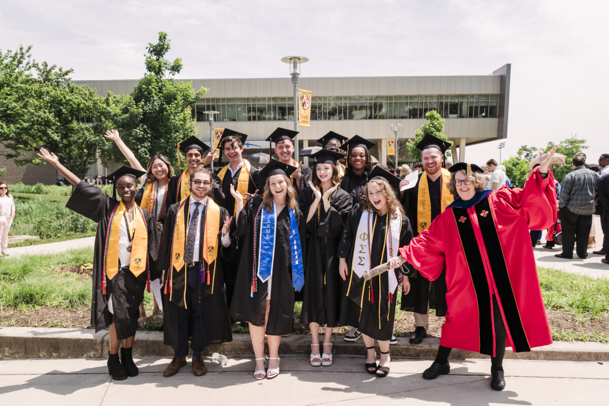 UMBC Class of 2024 graduates posing with faculty member outside on UMBC campus. Many of the people in the photo have their hands up
