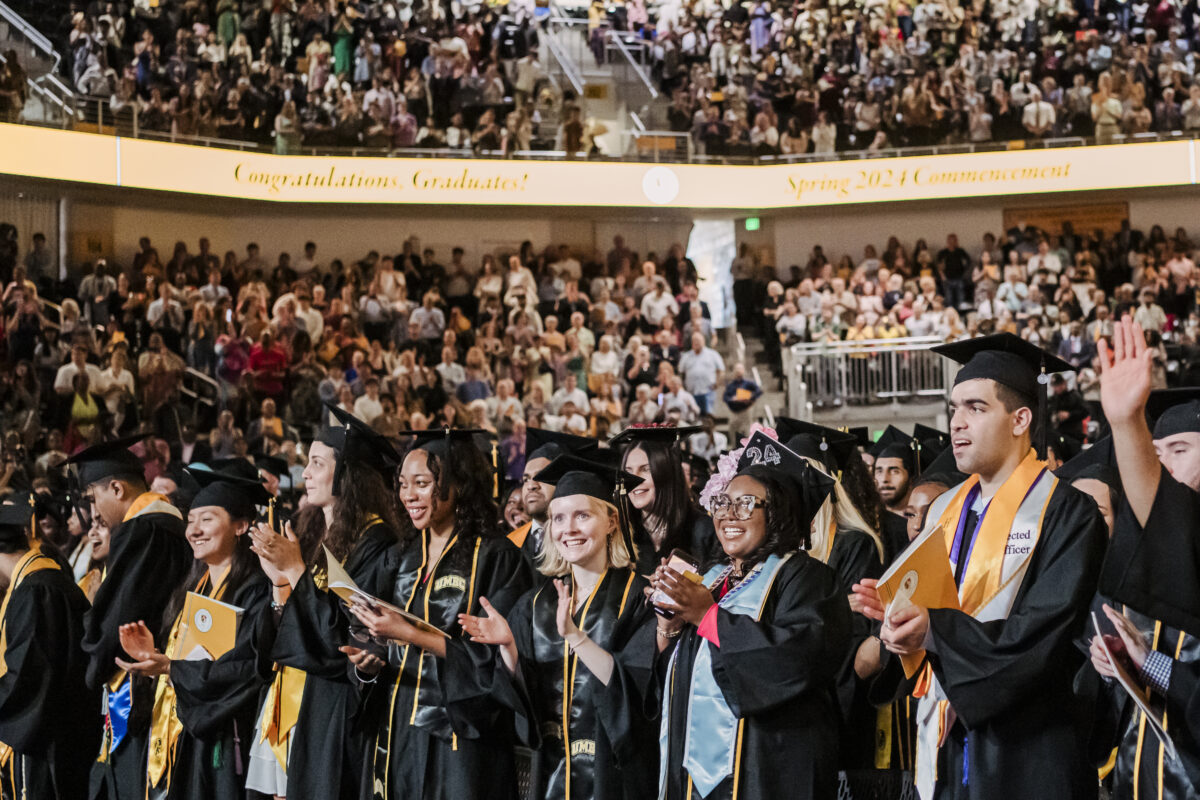 UMBC Class of 2024 graduates standing in the audience applauding and smiling. 