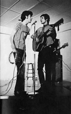 Two men on stage singing into a microphone. One playing tambourine, the other playing the guitar.