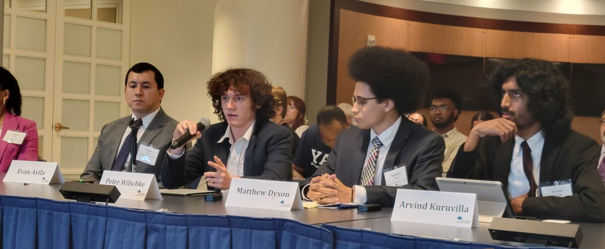 A group of four college students sit at a table, one is speaking into a microphone, presenting information with an audience behind them.