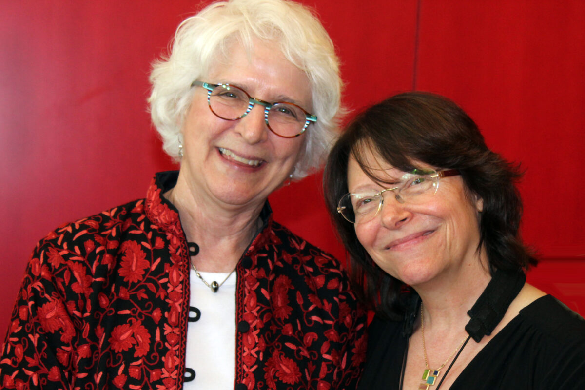 Two women, positioned against a bright red wall, smile at the camera. On the left, a woman with white hair, glasses, and red top; on the right a woman with dark hair, glasses, and a dark top.
