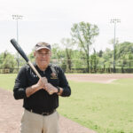 an older man in a black polo shirt stands on a baseball field holding a bat
