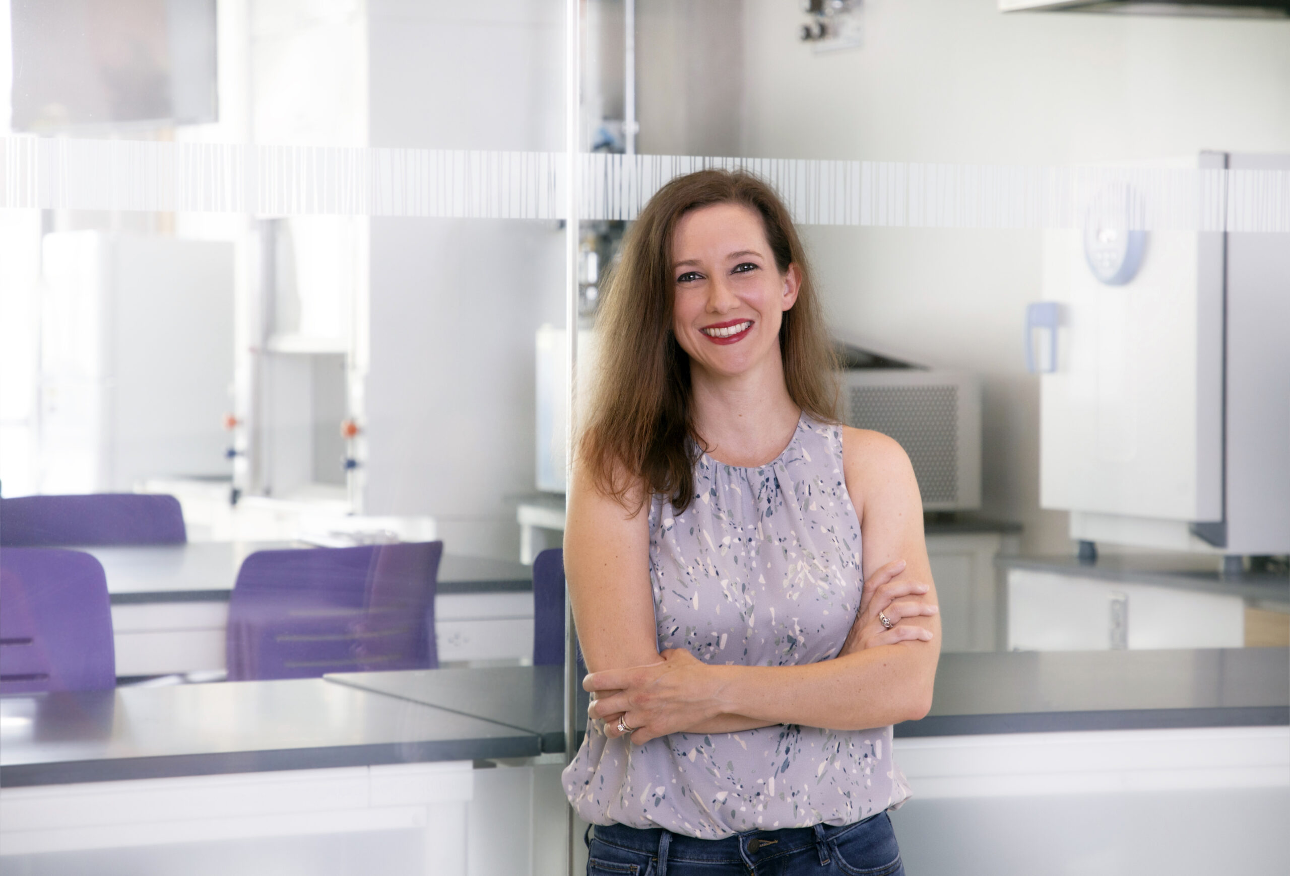 woman stands in front of glass wall, lab benches with purple chairs behind.