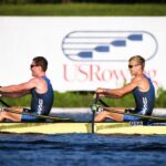 Two men in a rowing boat that is in water paddling oars in front of a backdrop that says USRowing