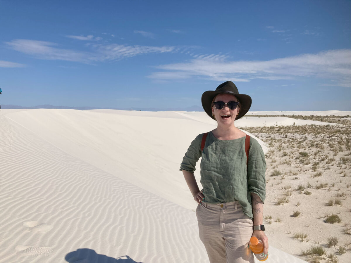 woman in sunglasses and broad-brimmed hat stands in huge field of sand, bright blue sky in background
