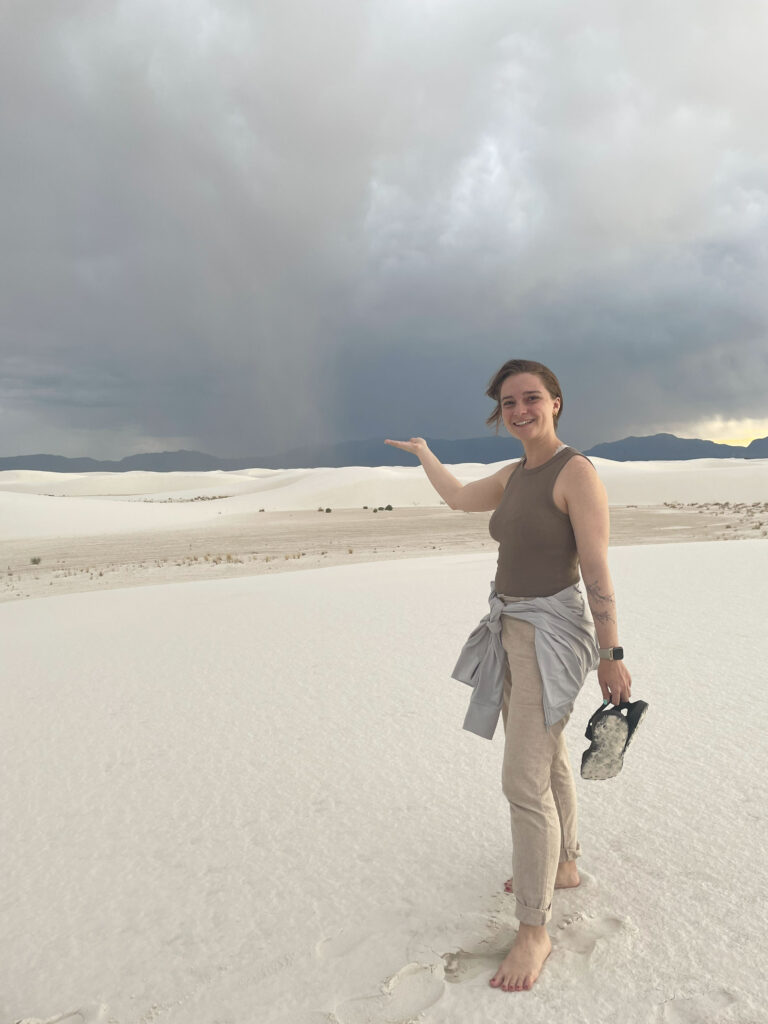 woman stands barefoot on sand and gestures toward a swirling cloud of dust in the distance