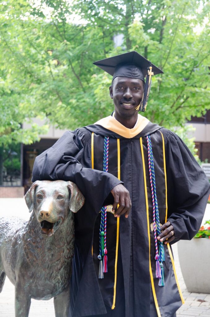 A college student wearing his cap and gown stands outside next to a statue of a dog