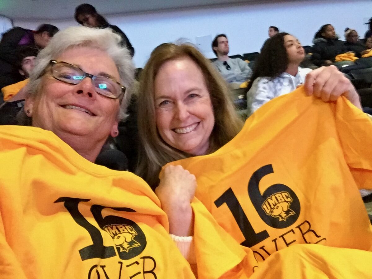two women hold up shirts commemorating UMBC's mens basketball win over UVA in 2018