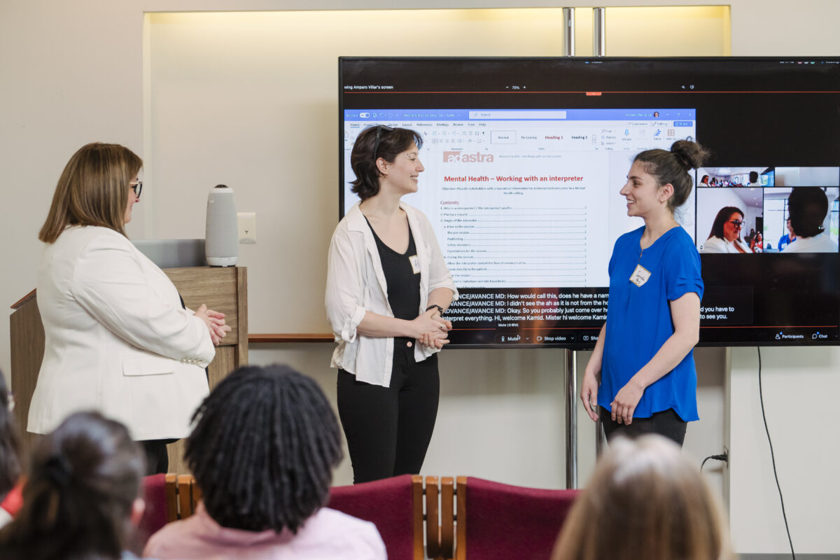 Two college students face each other while taking direction from a trainer in front of a large TV screen projecting a worksheet on working with a mental health interpreter