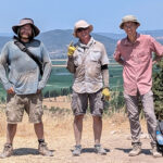 Three adults wearing dusty cloths stand on a clearing in Greece with mountains in the background