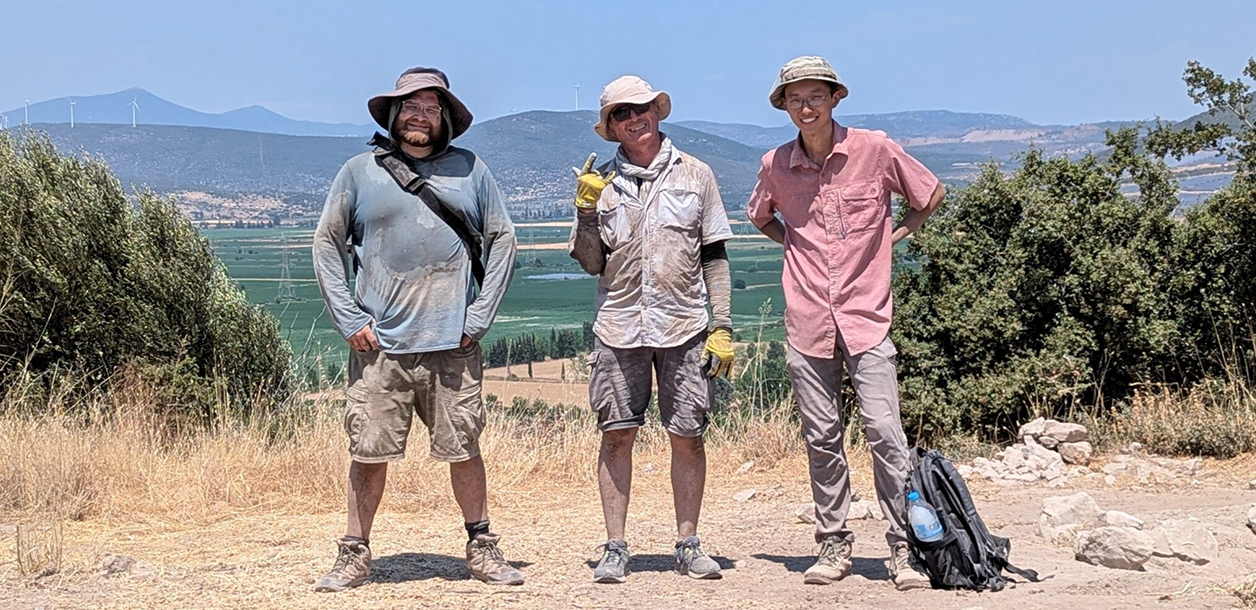 Three adults wearing dusty cloths stand on a clearing in Greece with mountains in the background