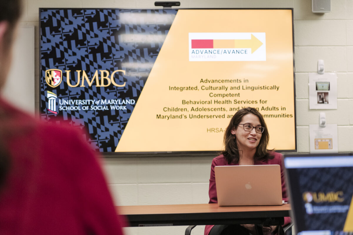 A professor sits at a table in front of a lap top with a projection screen behind them displaying the logos for University of Maryland Baltimore County, University of Maryland School of Social Work, and a new grant named ADVANCE or in Spanish AVANCE.