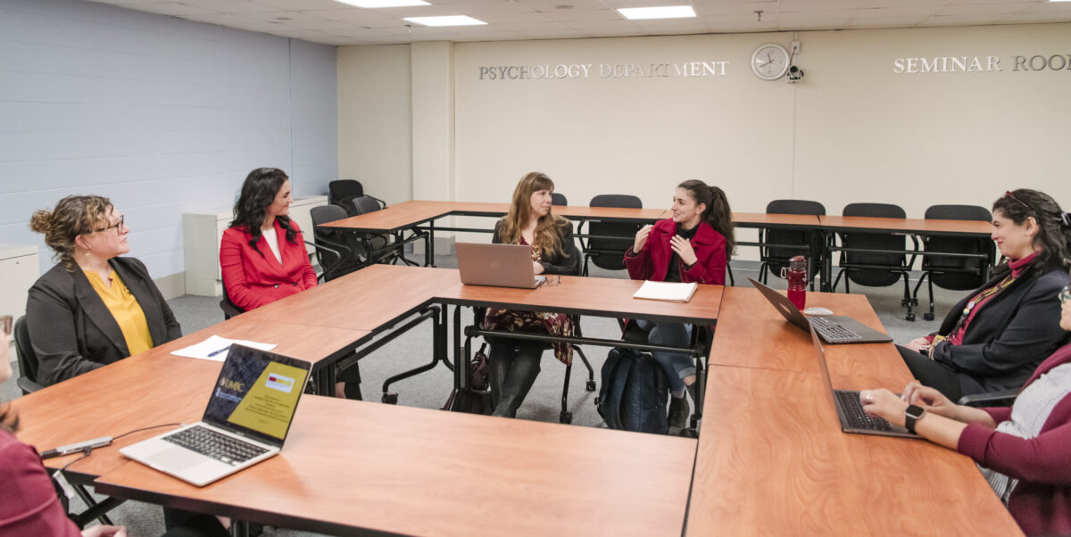 A graduate student with long black hair in a pony tail wearing a red coat sits with professors at tables in a classroom with a silver sign spelling psychology department talking about behavioral health in Maryland