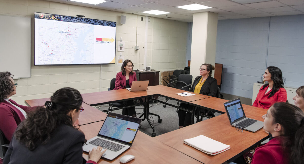 A group of professors and graduate students sit around tables arranged in a square and look at a projection screen that shows a map of Maryland with red dots for mental health training sites