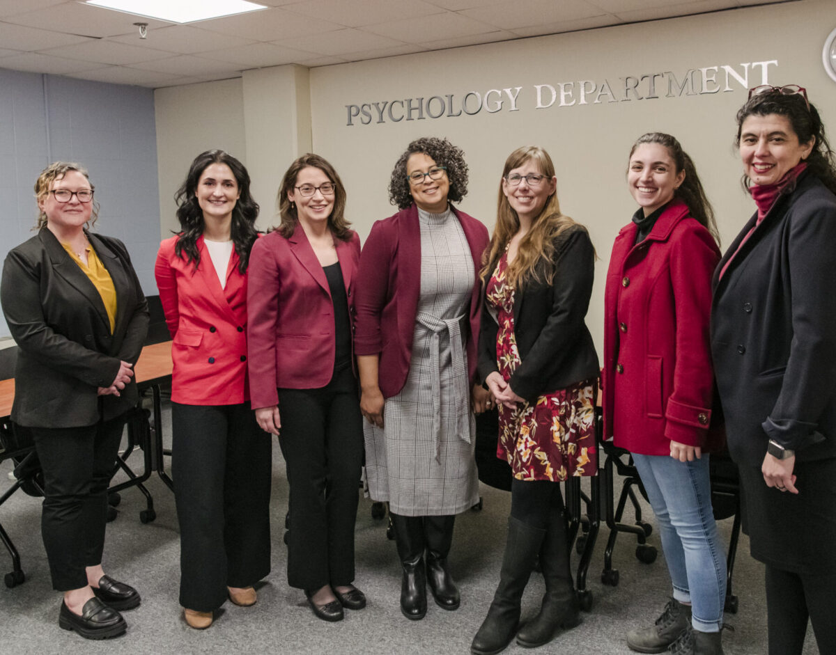 A group of seven made up of behavioral health psychology professors and graduate students stand side by side to take a picture inside a classroom with silver letters on the back wall spelling psychology department
