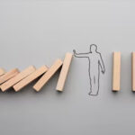 A line of wooden dominos, with a sketch of a person in the middle. The dominos on the left are falling, the person puts their hand up to stop them from hitting the dominos on the right.