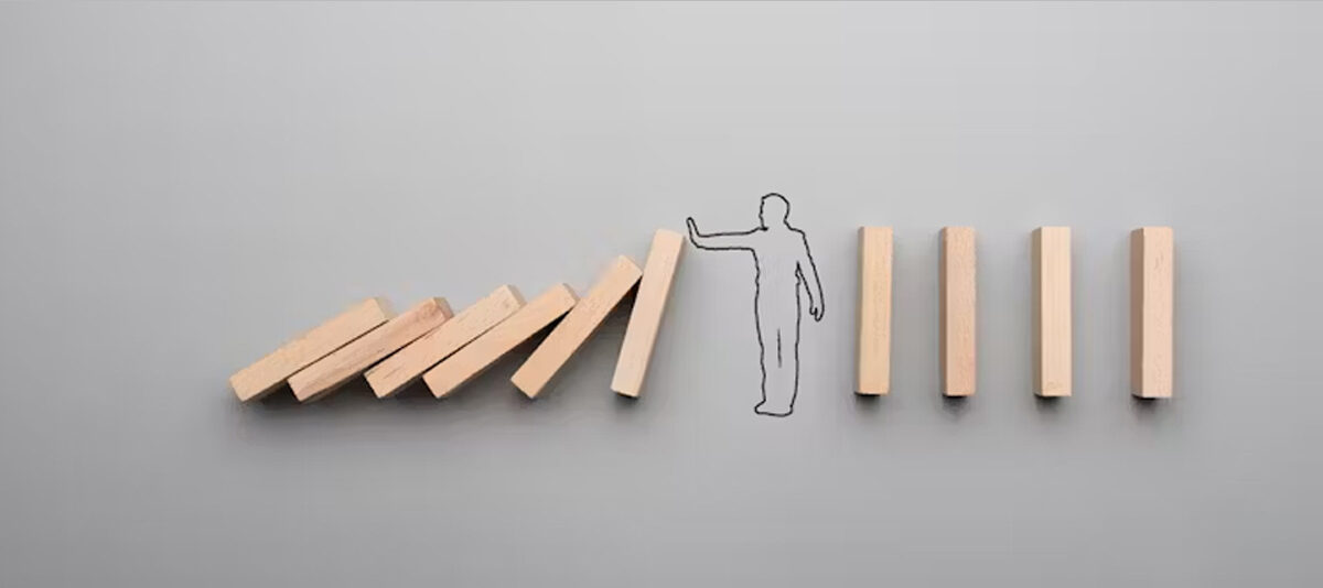 A line of wooden dominos, with a sketch of a person in the middle. The dominos on the left are falling, the person puts their hand up to stop them from hitting the dominos on the right.