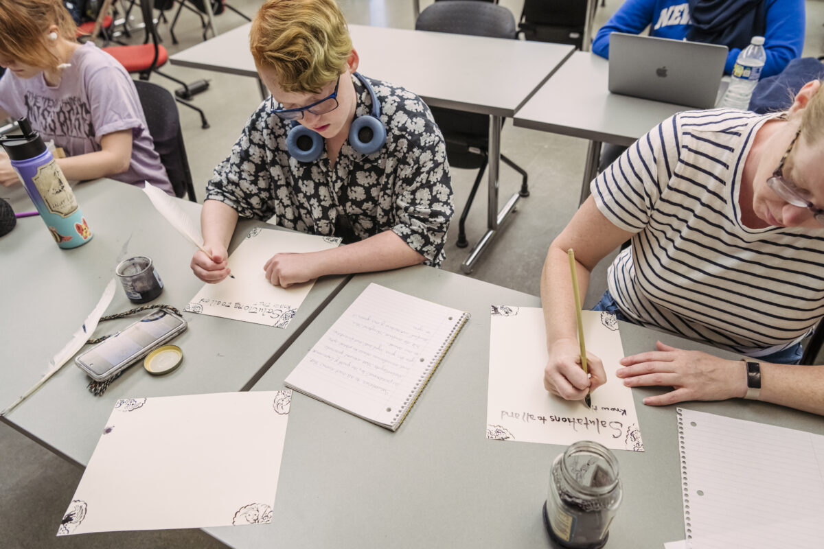 two students write with quill pens at a table