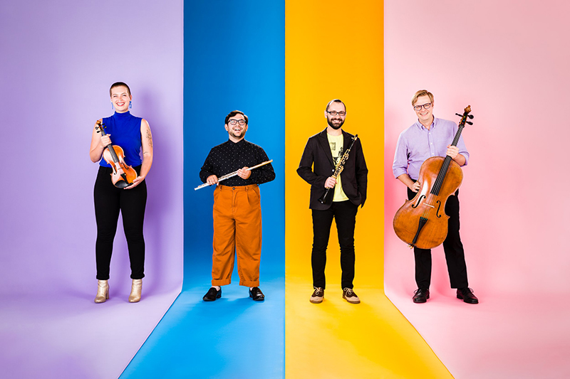 Against a colorful backdrop, four musicians pose with instruments.