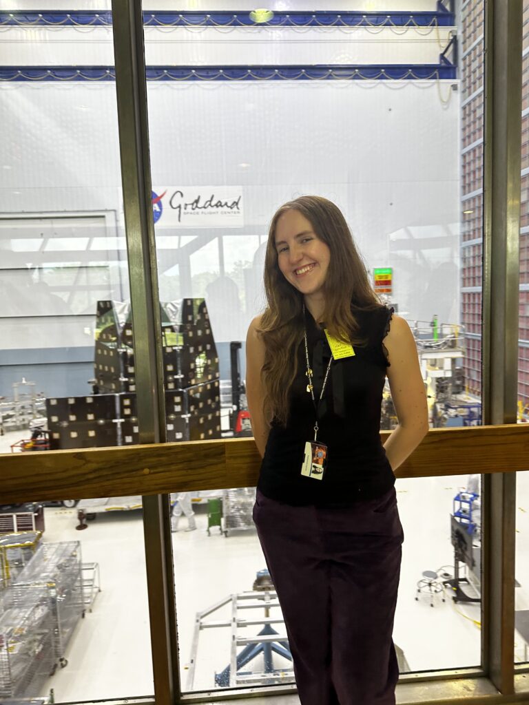 young woman stands in front of glass windows; behind the windows is a clean room, a science construction area of sorts, containing gold and black panels and other equipment