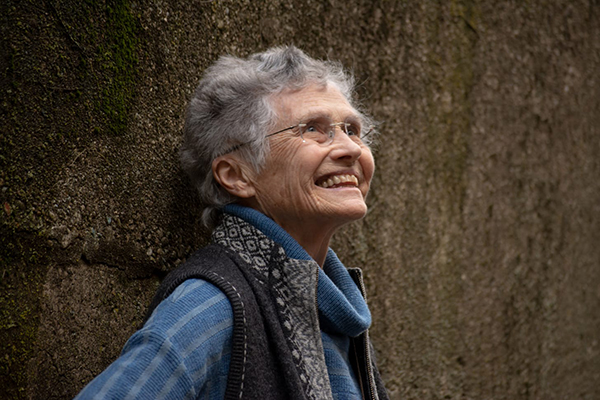 A person with glasses and grey hair, wearing a blue and black vest, smiles and looks upwards.