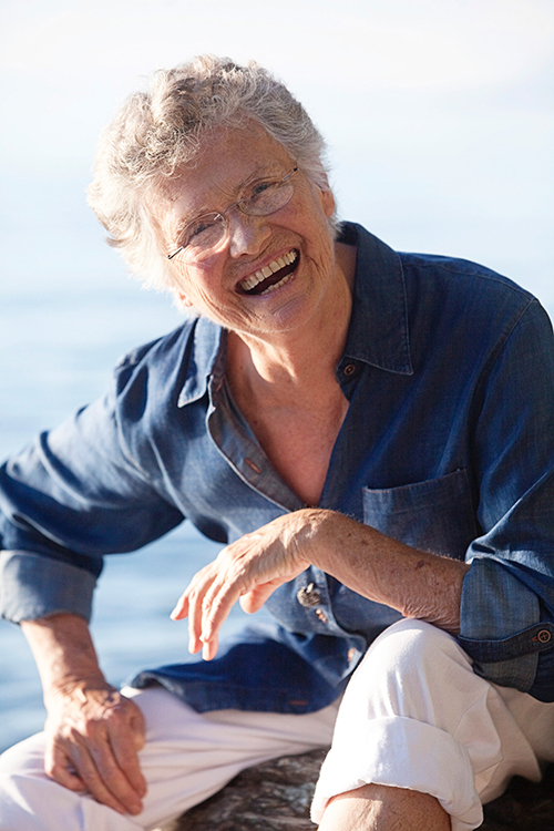 A person with gray hair and glasses, wearing a blue top and white pants, smiles at the camera.