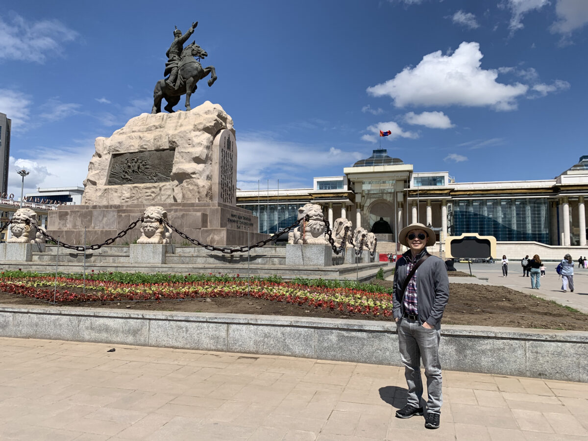 A researcher stand in front of large statue of a man on a horse sits on top of a large boulder with a plaque at the center of a plaza in Mongolia
