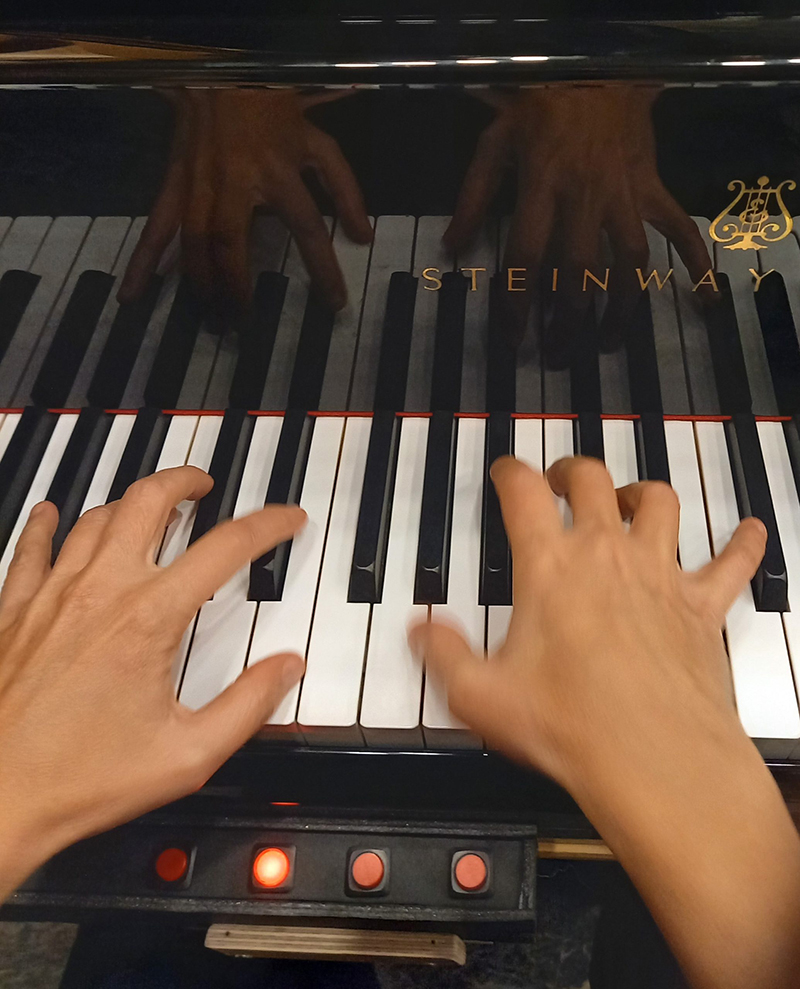 Two hands at a piano keyboard, a row of red buttons in front.